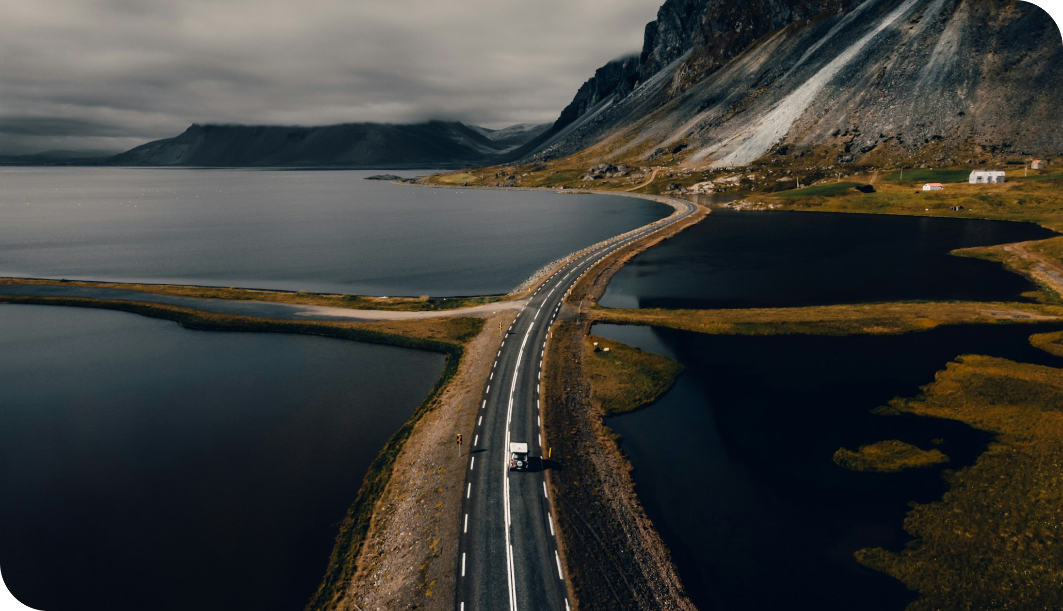Dramatic coastal road winding between calm lakes and rugged mountains under a cloudy sky in Iceland, with a lone vehicle travelling along a scenic remote landscape.